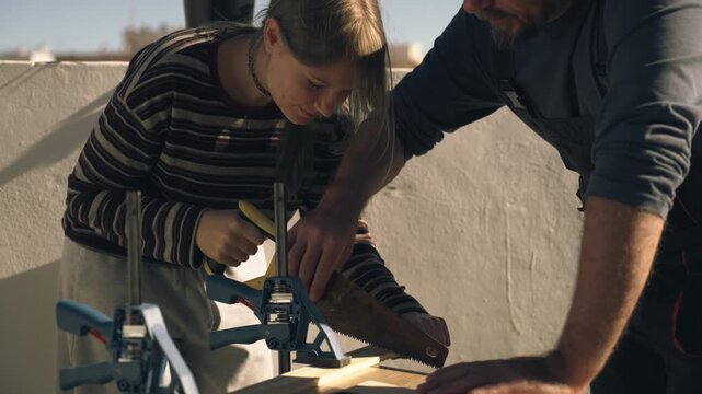 Young girl learning woodworking skills from father, skilled carpenter, as they saw wooden plank together on sunny terrace during home improvement project