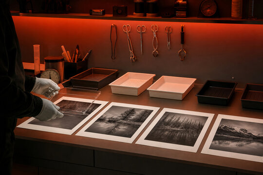 A person stands at a table in a darkroom looking at black and white prints. Various printing tools and trays are arranged on the table. The workspace is lit with a warm light.