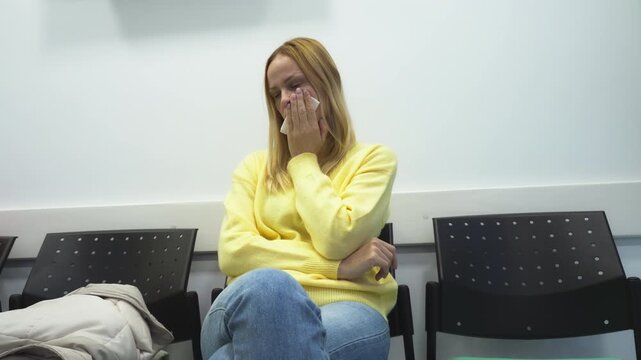 Blonde woman suffering severe toothache sitting in dental waiting room, holding tissue to her cheek and grimacing in pain while waiting for appointment in modern dental clinic environment.