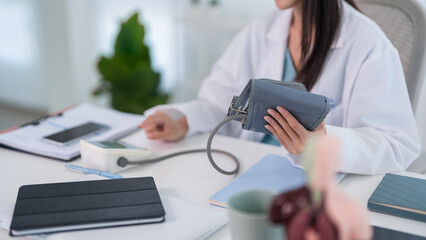 Fototapeta premium Asian adult female doctor measuring blood pressure with device at medical clinic office desk for health checkup examination cardiovascular hypertension