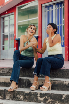 Dos j&oacute;venes amigas sonriendo y tomadas de la mano sentadas en unas escaleras urbanas celebrando el d&iacute;a de la juventud.
