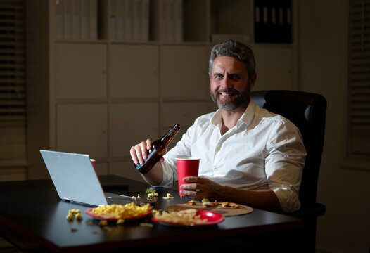 Freelancer working late with snacks in night home office. Business man night routine with laptop. Snack break during laptop work in office. Man at desk eating popcorn in late hours.