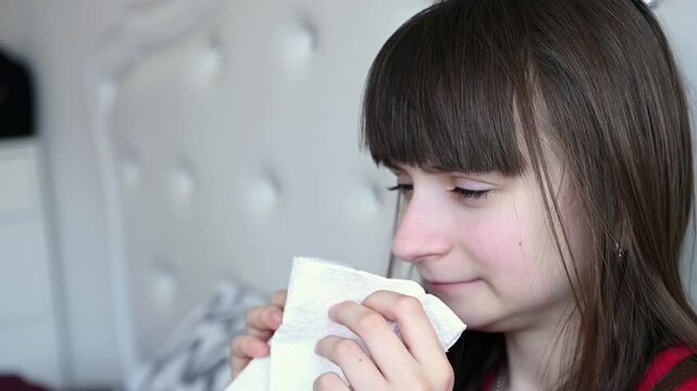 A teenage girl sits in bed and blows her nose into a paper napkin. Cold, runny nose, seasonal allergy.