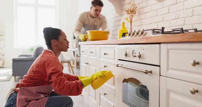 Couple cleaning kitchen with gloves showing teamwork. Man wipes oven while woman scrubs cabinets, bright home interior with bucket and sponge for hygiene. Shared home tasks show duty.