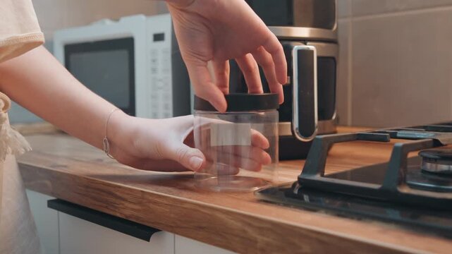 Jar beside stainless air fryer cooking, modern kitchen scene, hand positions transparent container on wooden counter near microwave and gas burner, preparing ingredients for quick healthy meal, warm