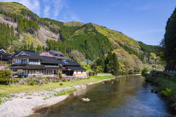 新緑の山々を背景に、澄んだ川のほとりに黒い瓦屋根の伝統的な日本家屋が建つ田舎の風景 © ToYoPHoTo