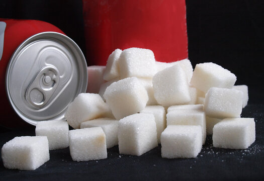 Unhealthy diet concept: A large pile of white sugar cubes next to generic red soda cans representing high calorie and sugar content in soft drinks