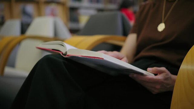 A woman sits in a comfortable chair in a library or bookstore, holding a book open on her lap and turning the pages.