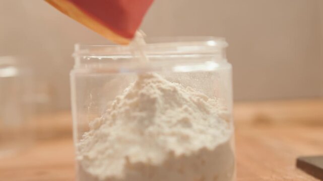 Closeup of flour being transferred into jar. Warm wooden surface highlights careful flour scooping process. Detailed view of baker pouring fine flour into glass container on wooden counter