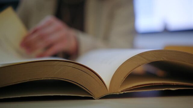 Close-up shot of an open book on a desk. A woman in the background is reading and turning the pages, studying or researching in a library.