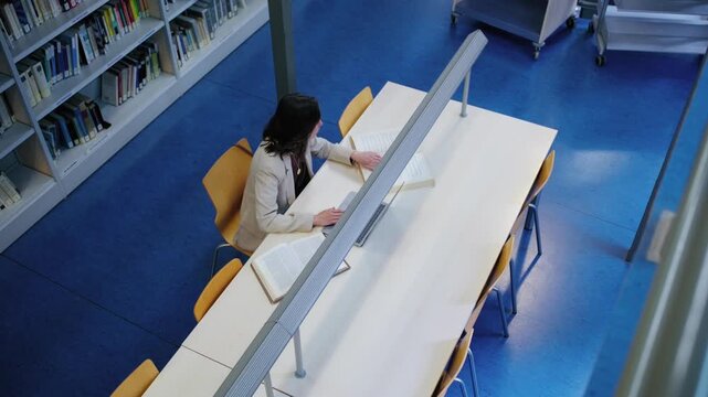 A woman studies with a laptop and books at a table in a modern university library. High angle view of a student researching and learning.