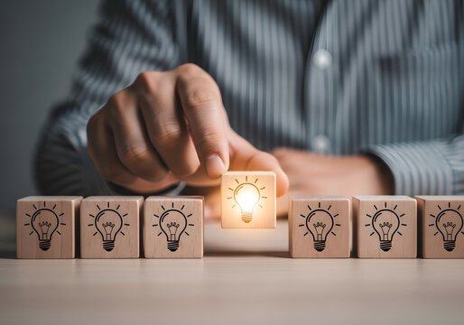 A person's hand placing a luminous lightbulb cube amongst identical unlit cubes, symbolizing the moment of inspiration, conceptual breakthrough, and the genesis of a brilliant new thought