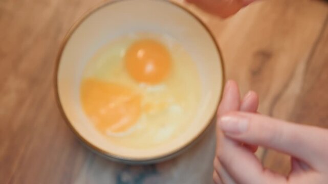 Hands cracking egg into bowl gently over ceramic mixing bowl, fingers separate shell, yolk settles into dish, home cook practicing technique, warm wooden counter, morning routine for breakfast recipe