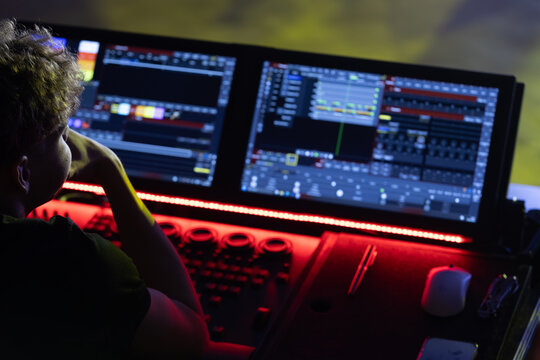 Individual with curly hair operates dual computer monitors displaying audio and visual software in a dimly lit control room with colorful lighting effects