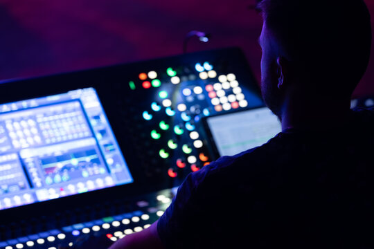 Male sound engineer operating a digital audio mixer with colorful buttons and screens in a dimly lit studio environment during a live performance setup
