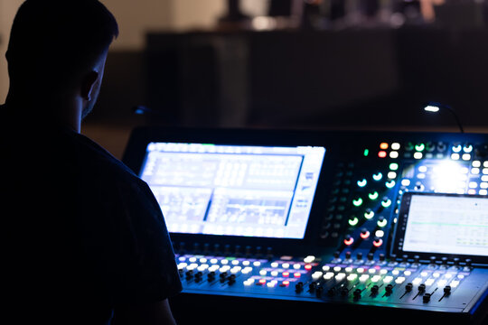 Sound technician operating a digital audio mixer with illuminated controls in a dimly lit venue, focused on the mixing process during a live performance setup