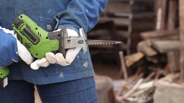 Gardener holding a cordless reciprocating saw to trim tree branches. Hand-held cordless saw for gardening. Close-up of hands in protective gloves holding a compact electric saw