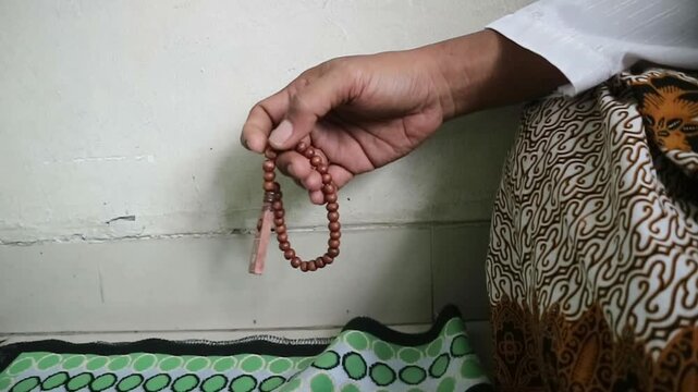 Close-up image of a Muslim man's hands reciting dhikr with wooden prayer beads while praying, wearing a traditional Javanese batik sarong.