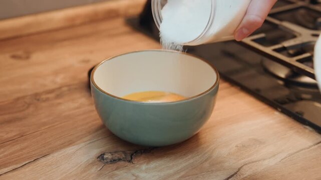 Measuring sugar over eggs bowl, steady hand pouring sugar stream into green ceramic bowl on modern countertop, instructional baking prep step, clean bright kitchen lighting, closeup focus on technique
