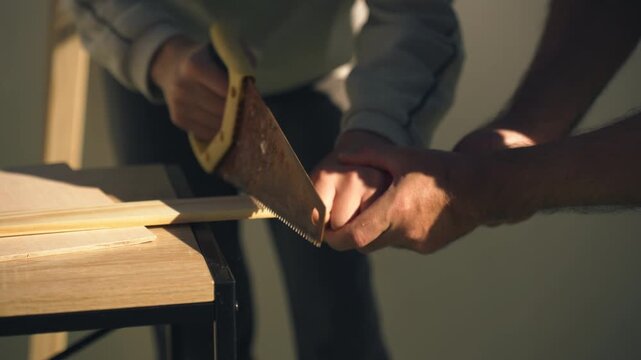 Close-up of father guiding his teenage son&rsquo;s hands while sawing a wooden plank together, demonstrating woodworking skills and hands-on training in a DIY home project.