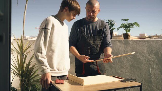 Close guidance from father teaching teenage son how to measure and cut wooden plank using tools, focusing on woodworking skills and hands-on training.