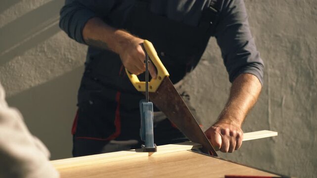 Close-up of male worker in uniform cutting wooden board with hand saw in outdoor setting, representing DIY home improvement and practical woodworking skills.
