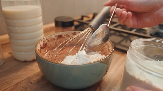 Measuring flour for cocoa batter, pastry chef mimics precise spooning technique into chocolate base, whisk on counter, milk bottle and glass jar in background, instructional closeup for recipe content