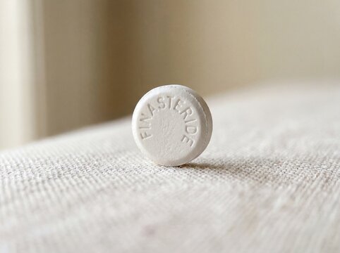 A professional photography close-up shot of a small round white pill with FINASTERIDE engraved on the top, placed on a soft neutral background.