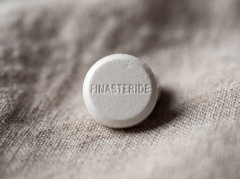 A professional photography close-up shot of a small round white pill with FINASTERIDE engraved on the top, placed on a soft neutral background.
