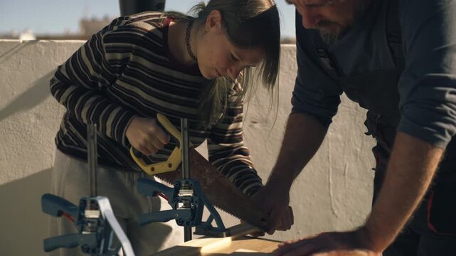 Father teaching teenage daughter how to use a hand saw properly while cutting wood outdoors, demonstrating basic construction skills.