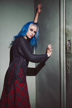 Indoor portrait of cool grunge (rock) girl. Informal blue-haired model, dressed in checkered skirt, and holey sweater standing at the wall