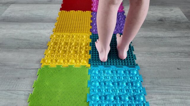 In horizontal slow-motion video, a child steps onto colorful rubber massage mats, engaging in autonomous foot care exercises to prevent common pediatric foot issues. Early intervention program clips