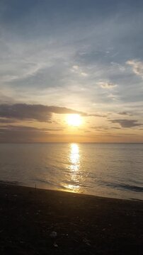 Beautiful golden sunset over a black sand beach with calm sea water and colorful evening sky. Peaceful coastal scenery with warm sunlight reflection, located at Singaraja, Bali, Indonesia.