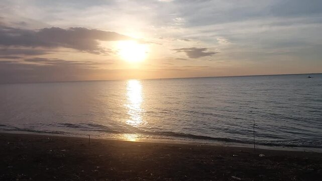 Beautiful golden sunset over a black sand beach with calm sea water and colorful evening sky. Peaceful coastal scenery with warm sunlight reflection, located at Singaraja, Bali, Indonesia.