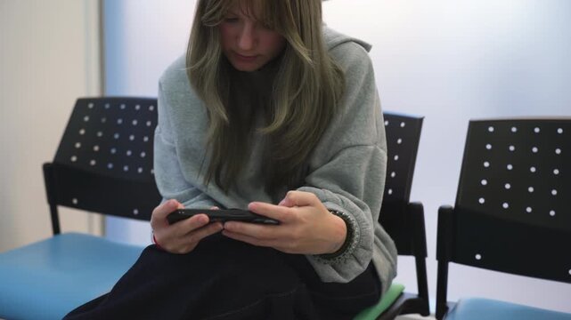 Teenage girl sitting alone on chair in public waiting area using smartphone, highlighting isolation, waiting time, and digital engagement in modern everyday life