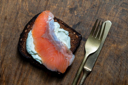 Open rye bread sandwich with cream cheese and smoked salmon slice served with golden cutlery on dark wooden table, closeup, top view