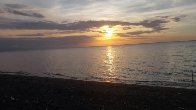 Beautiful golden sunset over a black sand beach with calm sea water and colorful evening sky. Peaceful coastal scenery with warm sunlight reflection, located at Singaraja, Bali, Indonesia.