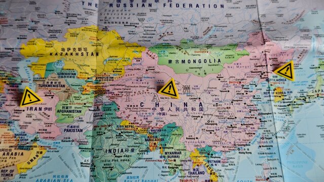 Yellow exclamation warning signs are placed over Iran, China, and North Korea on an Asian map, indicating regional hot spots and geopolitical risks.