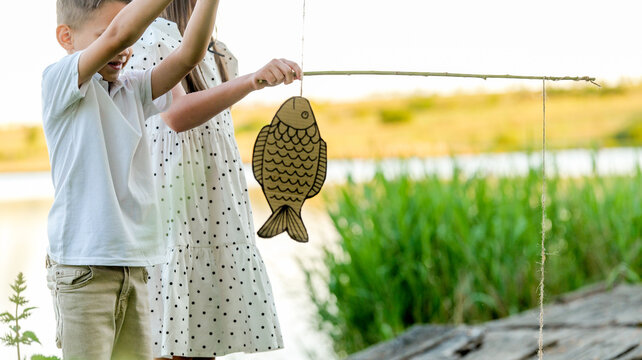 Children pretend fishing with a cardboard fish and a stick by the water