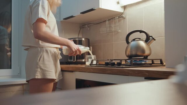 Sunlit kitchen pouring milk beside kettle morning breakfast prep with milk carton and coffee maker on countertop, casual loungewear, steam from kettle, inviting cozy atmosphere and simple routine.