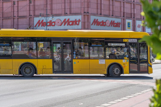 Germany, Berlin, May 25, 2026 A yellow Berlin bus passes the MediaMarkt facade at Alexanderplatz. Passengers, reflections, shop signs and leafy trees shape the urban scene.