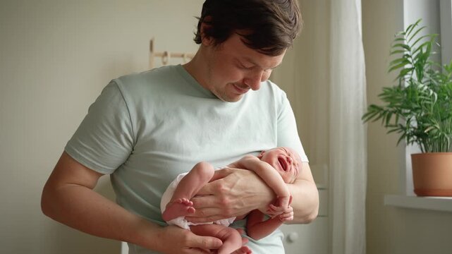 Young caucasian father gently rocking his newborn baby in his arms, trying to calm the crying infant. The intimate moment highlights paternal love, care, and the strong connection in a family