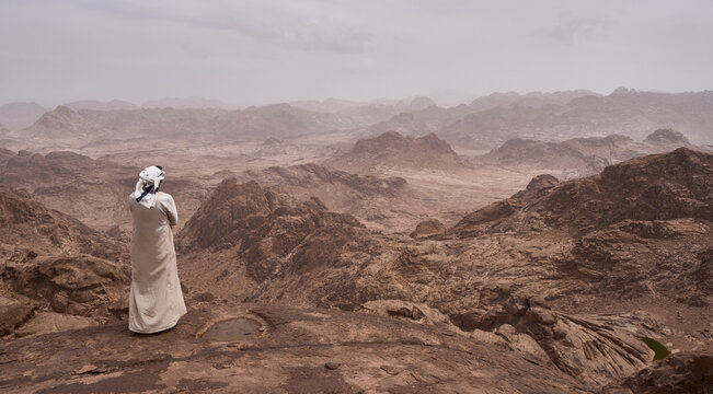 A bedouin in traditional clothing, long white robe with headscarf, in summit of Jebel Al‑Hashwa. Panorama of high mountains in national park Saint Katherine Protectorate. Authentic bedouin lifestyle.