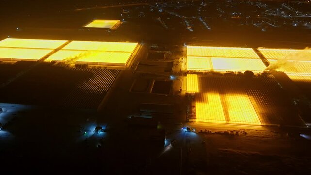 Aerial night panorama of illuminated greenhouses forming glowing patterns across a rural agricultural complex, contrasting with surrounding dark fields and distant lights