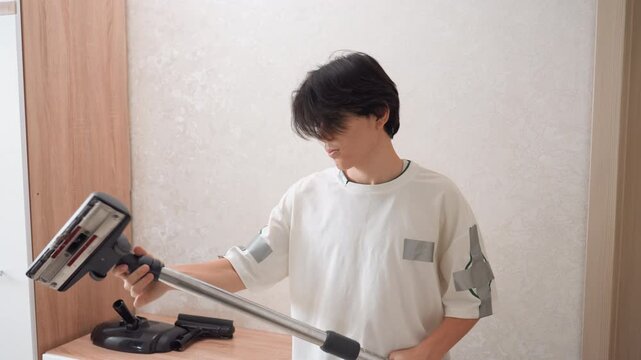 Young asian man assembling vacuum stick at home, testing cordless attachment and handle, focused expression, casual white shirt, modern minimal interior, preparing for house cleaning, stepbystep