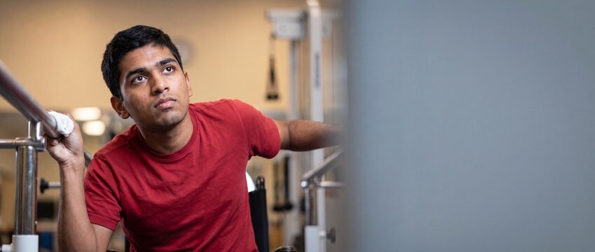 Disabled Indian man physical therapy rehabilitation exercising on parallel bars in clinical gym determined to walk again, hopeful disabled male patient recovery training studio background copy space.