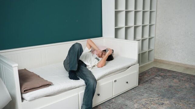 Woman relaxing on clean daybed in modern living room with teal accent wall, builtin shelving and patterned rug, storage bench with drawers, wearing jeans and tee while scrolling phone, calm cozy