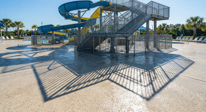 A metal staircase tower for a water slide complex casts strong geometric shadows onto the wet concrete deck of an outdoor water park on a sunny day.