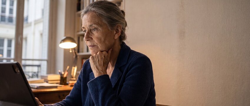Senior woman using tablet computer at home office desk, mature female with grey hair working remotely in cozy living room, concentrated thoughtful professional studying online, pensive digital lifesty