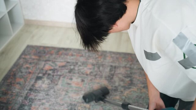 Young asian man spotcleaning rug at home, handheld vacuum attachment on patterned carpet, careful posture and focused technique, removing localized dirt with gentle motions in cozy room